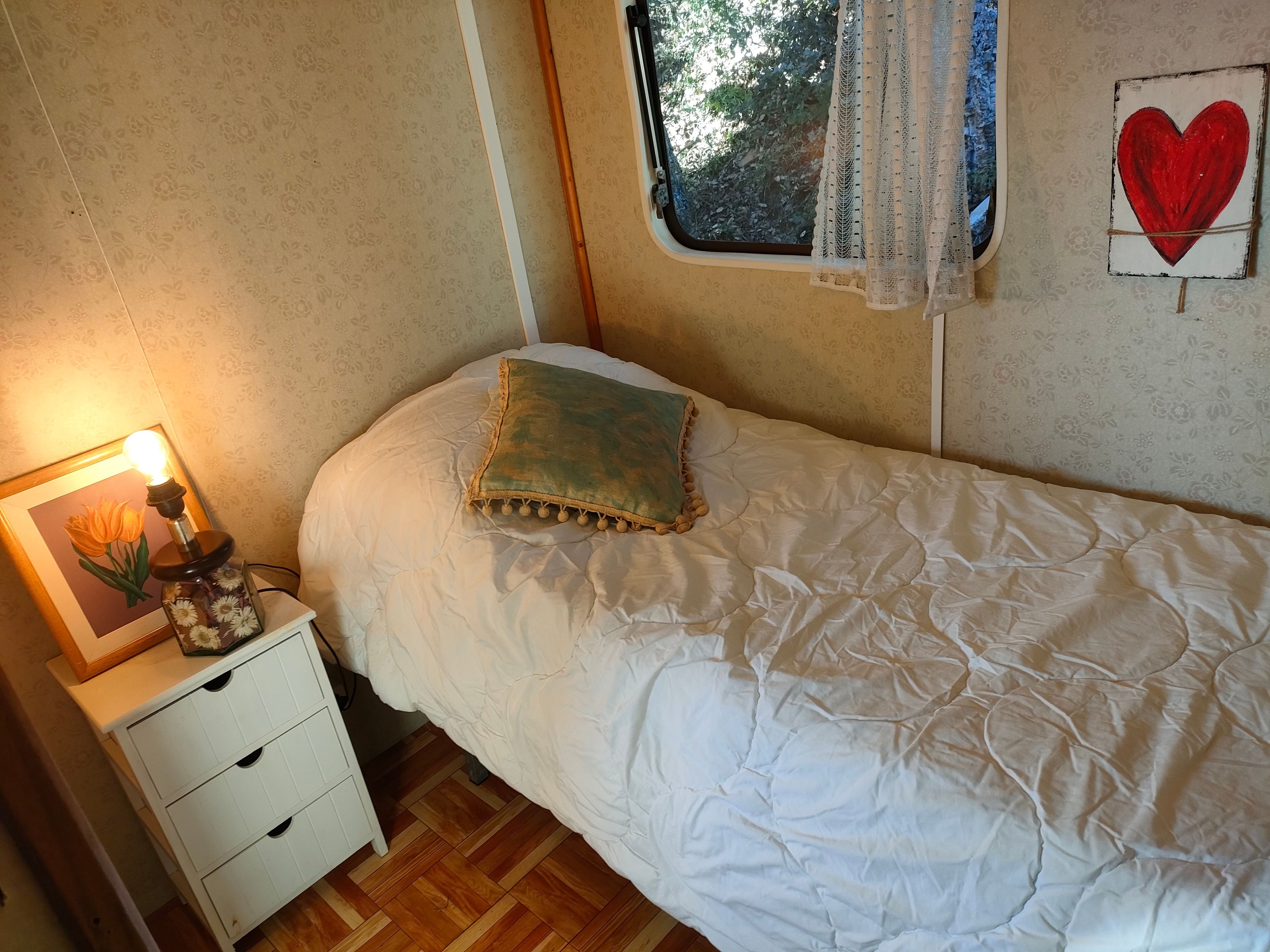 Une chambre à coucher avec un lit, une table de chevet avec une lampe, une fenêtre avec des rideaux et un élément décoratif mural.