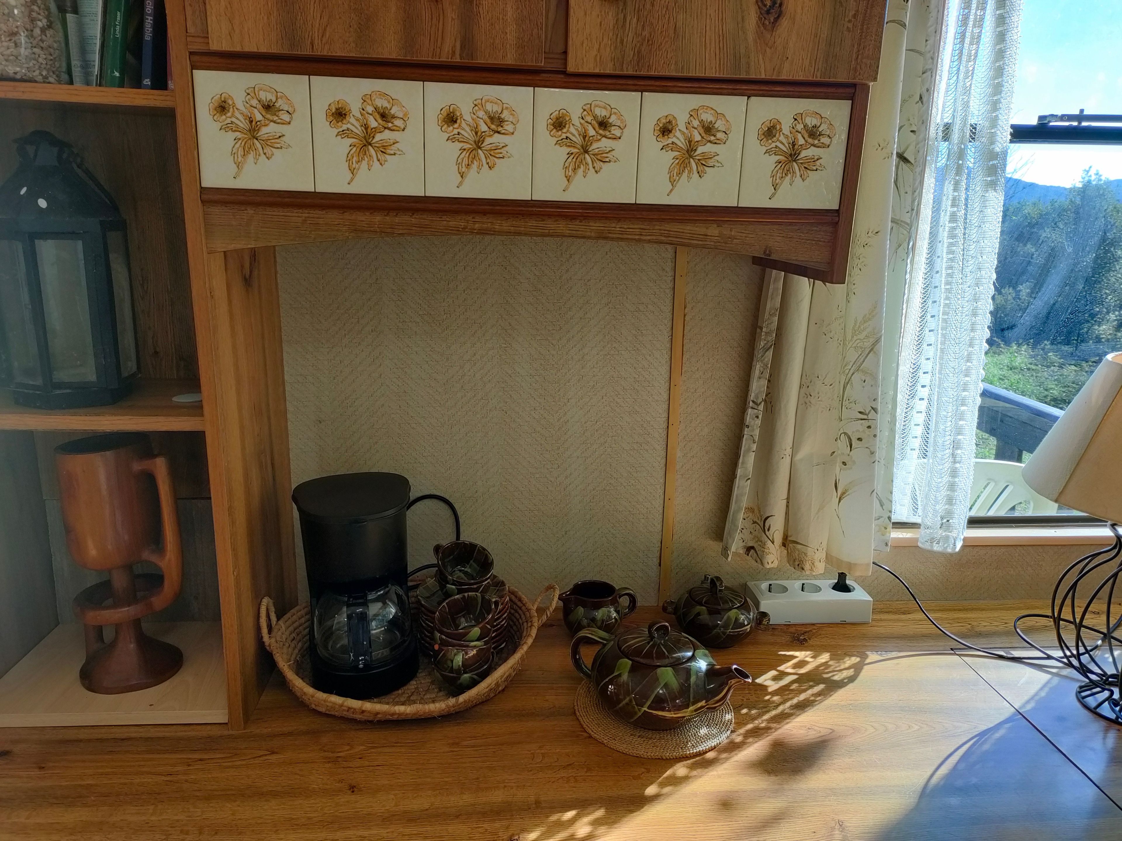 A wooden cabinet with a floral tile design, a bookshelf with books and a lantern, and a table with a teapot, cups, and a coffee maker.