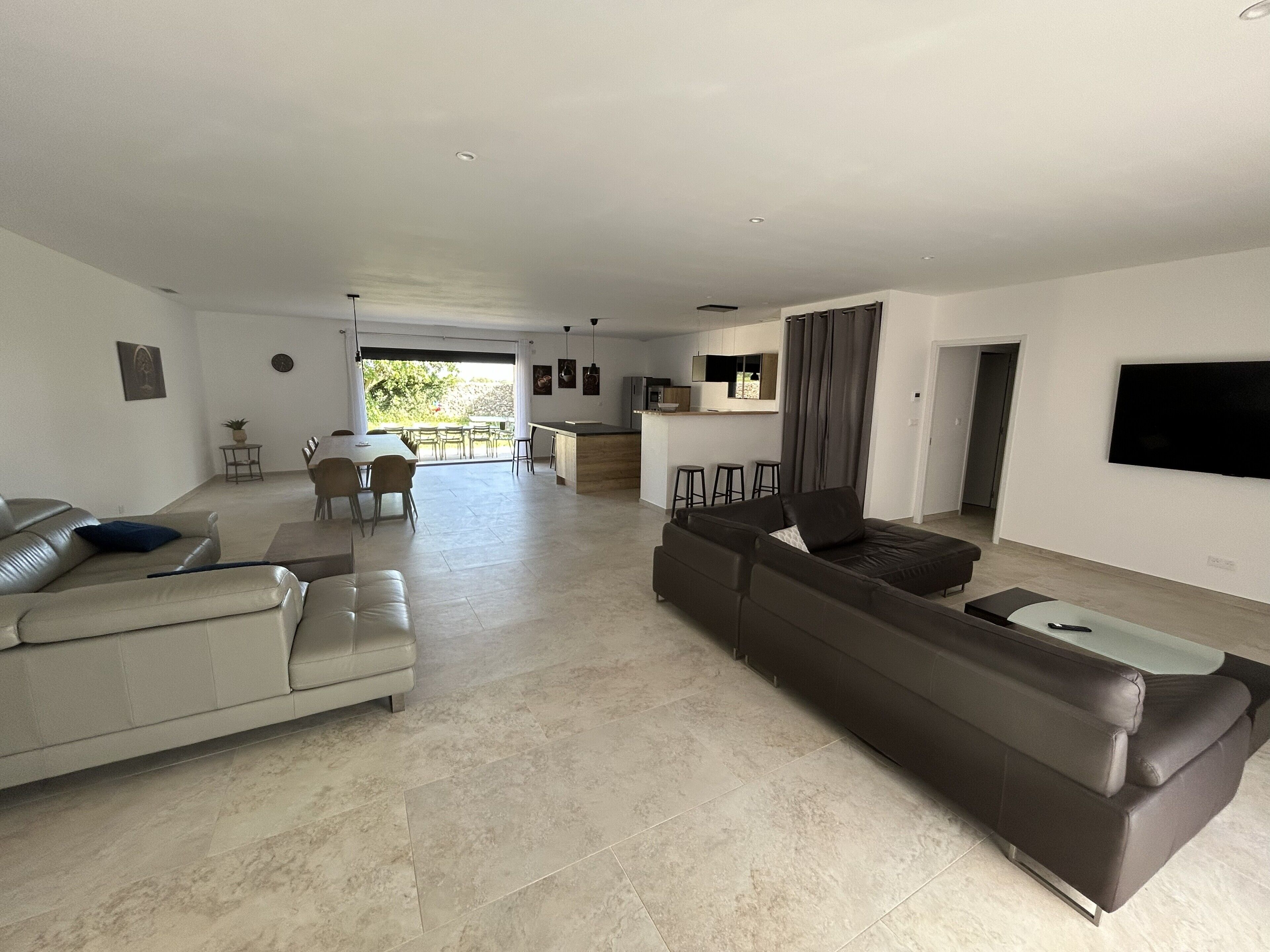A modern living room with a sofa, a dining area, a kitchen, and a TV mounted on the wall.