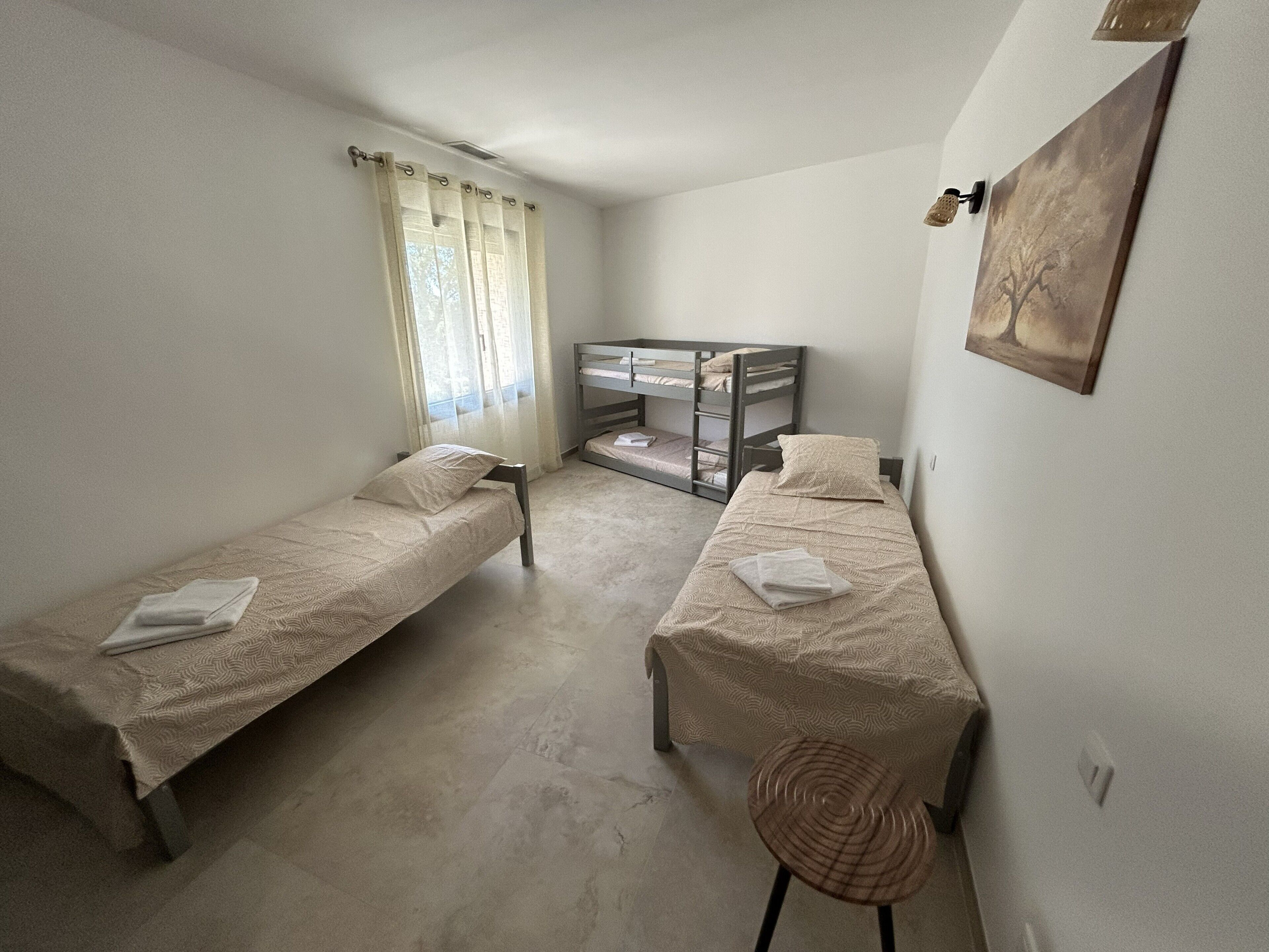 A small, clean room with two single beds, a bunk bed, a chair, and a window with curtains.
