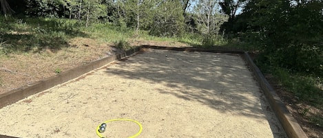 A sandy outdoor area with a metal hoop and a ball, surrounded by a wooden border and dense greenery.