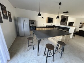 Eine moderne Küche mit einer zentralen Kochinsel, Barhocker, einem Kühlschrank, einem Backofen und Pendelleuchten.