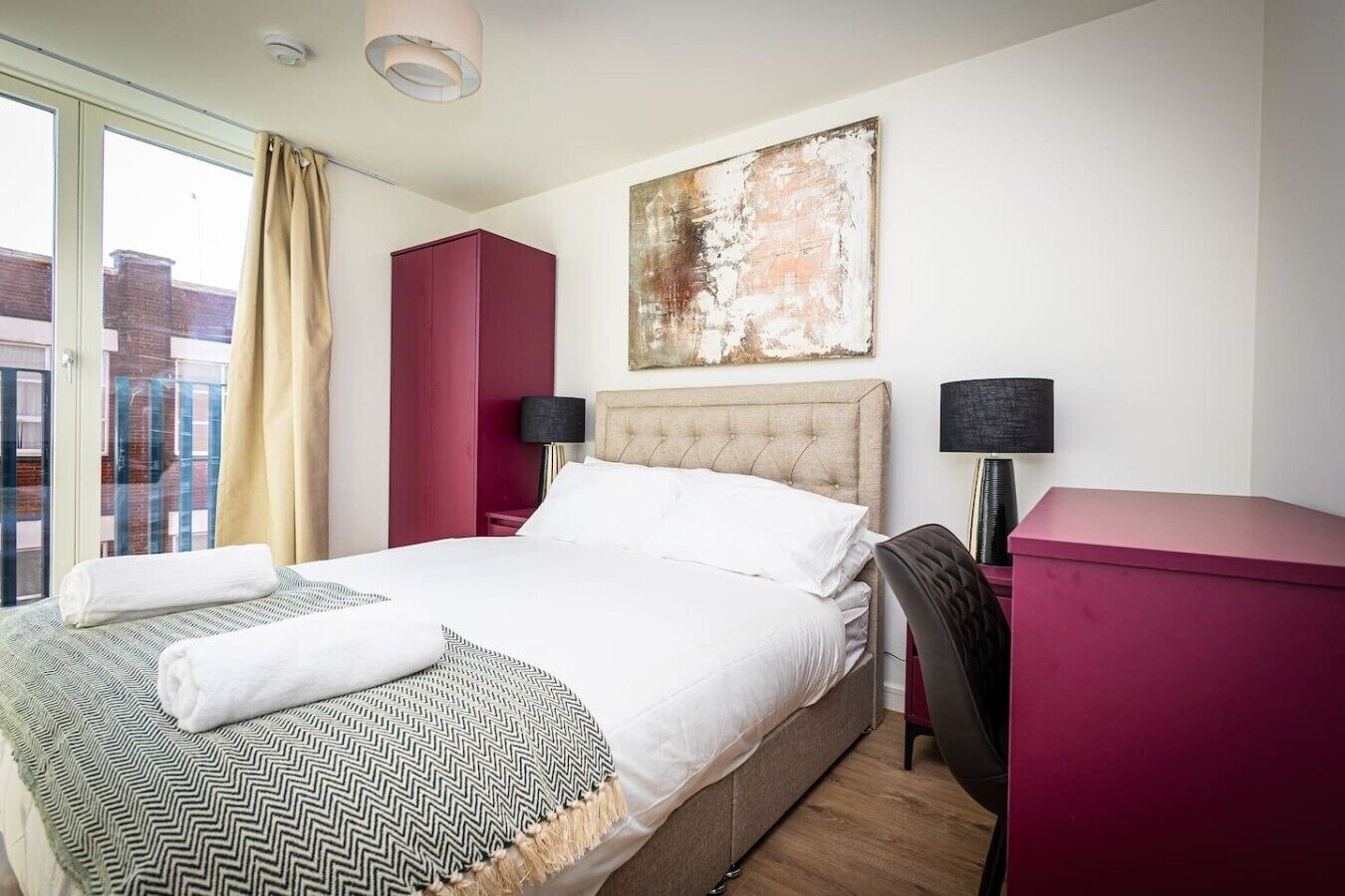 A hotel room with a bed, a desk, a chair, a red cabinet, a painting, and a view of the outside.