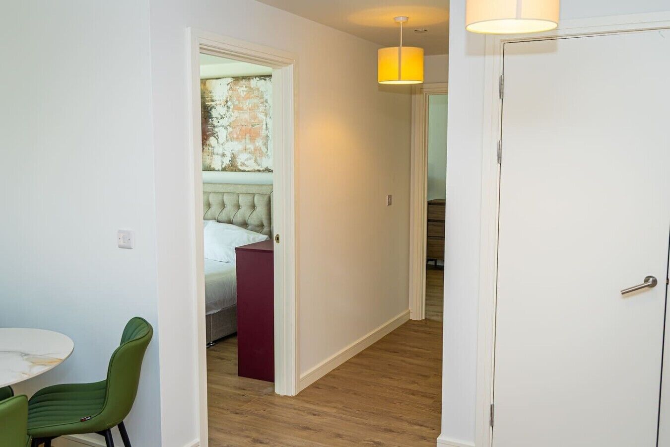 A hallway with a bed, a red nightstand, a dining area with a table and chairs, and a door.