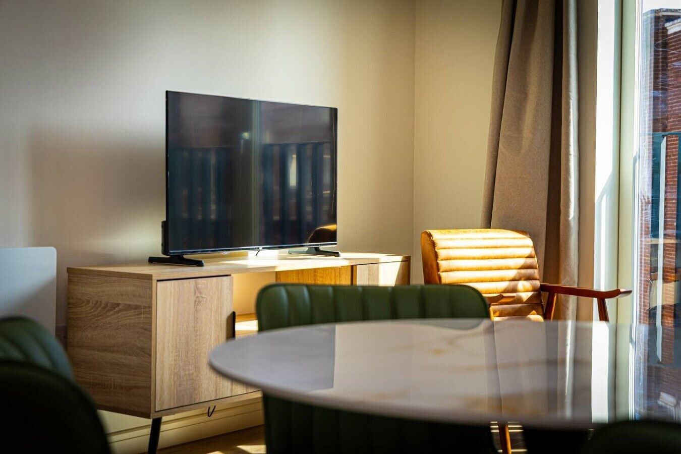 A hotel room with a flat-screen TV on a wooden cabinet, a round dining table with chairs, and a large window with curtains.