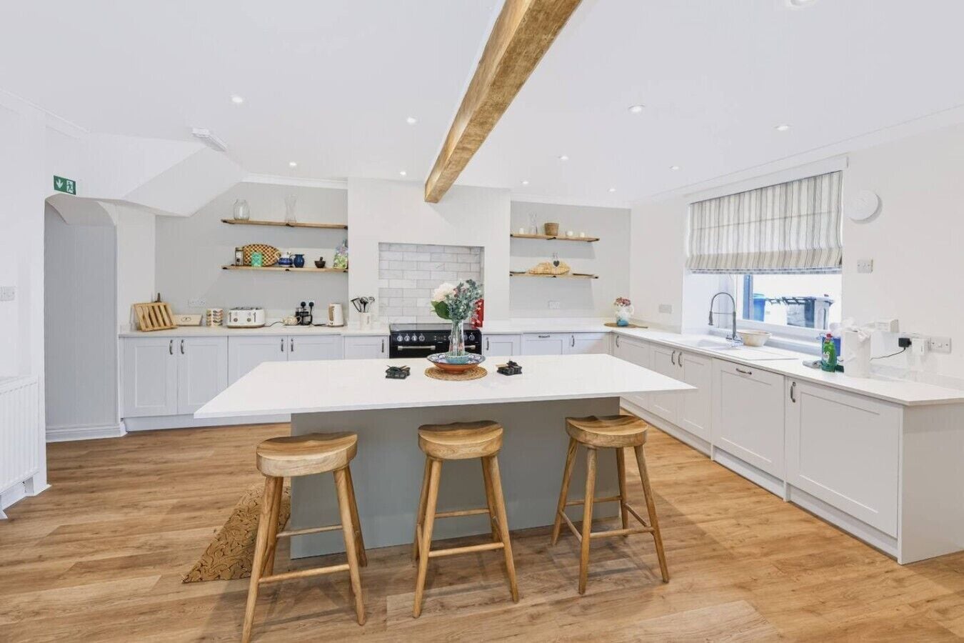 Une cuisine moderne avec des meubles blancs, un îlot central doté de tabourets, des poutres en bois au plafond et une fenêtre avec des stores.