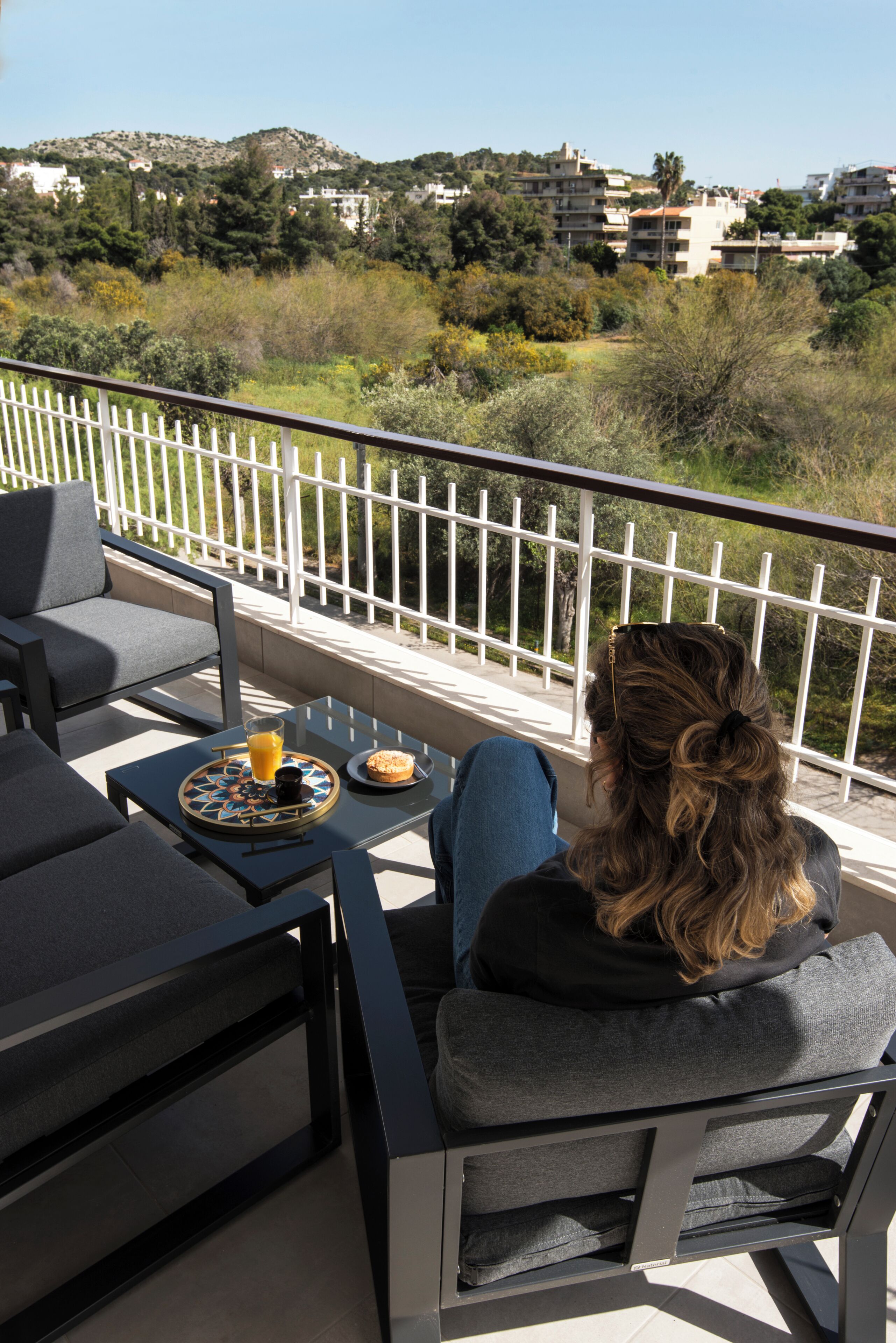 A balcony with outdoor furniture, a table with a glass of orange juice and a plate of food, and a view of a hillside with buildings.