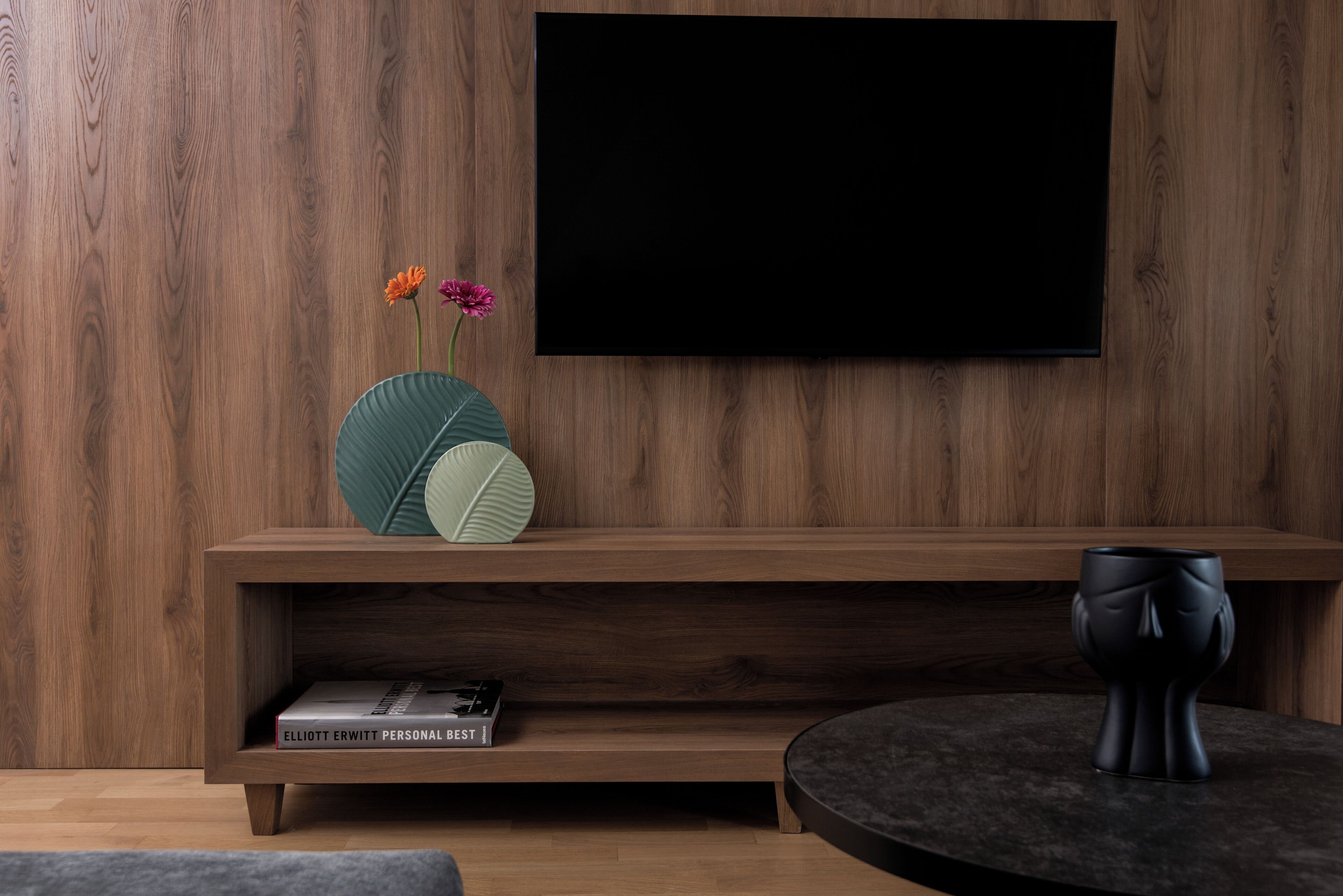 En moderne stue med en trævæg, et fladskærms-TV monteret over et trækonsolbord, en dekorativ vase med blomster, en bog med titlen 'Personal Best' og et rundt bord med en sort genstand på.