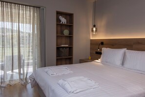 A hotel room with a bed, a nightstand, a lamp, a shelf with decorative items, and a window with curtains. - Krabo Luxury Suite (Vouliagmeni)
