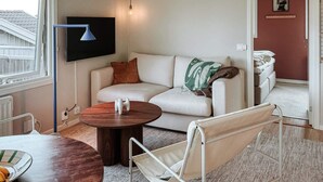 A modern living room with a sofa, a round wooden table, a TV mounted on the wall, and a window with a view of a house. - 5 Star Holiday Home in Uddevalla (Uddevalla)