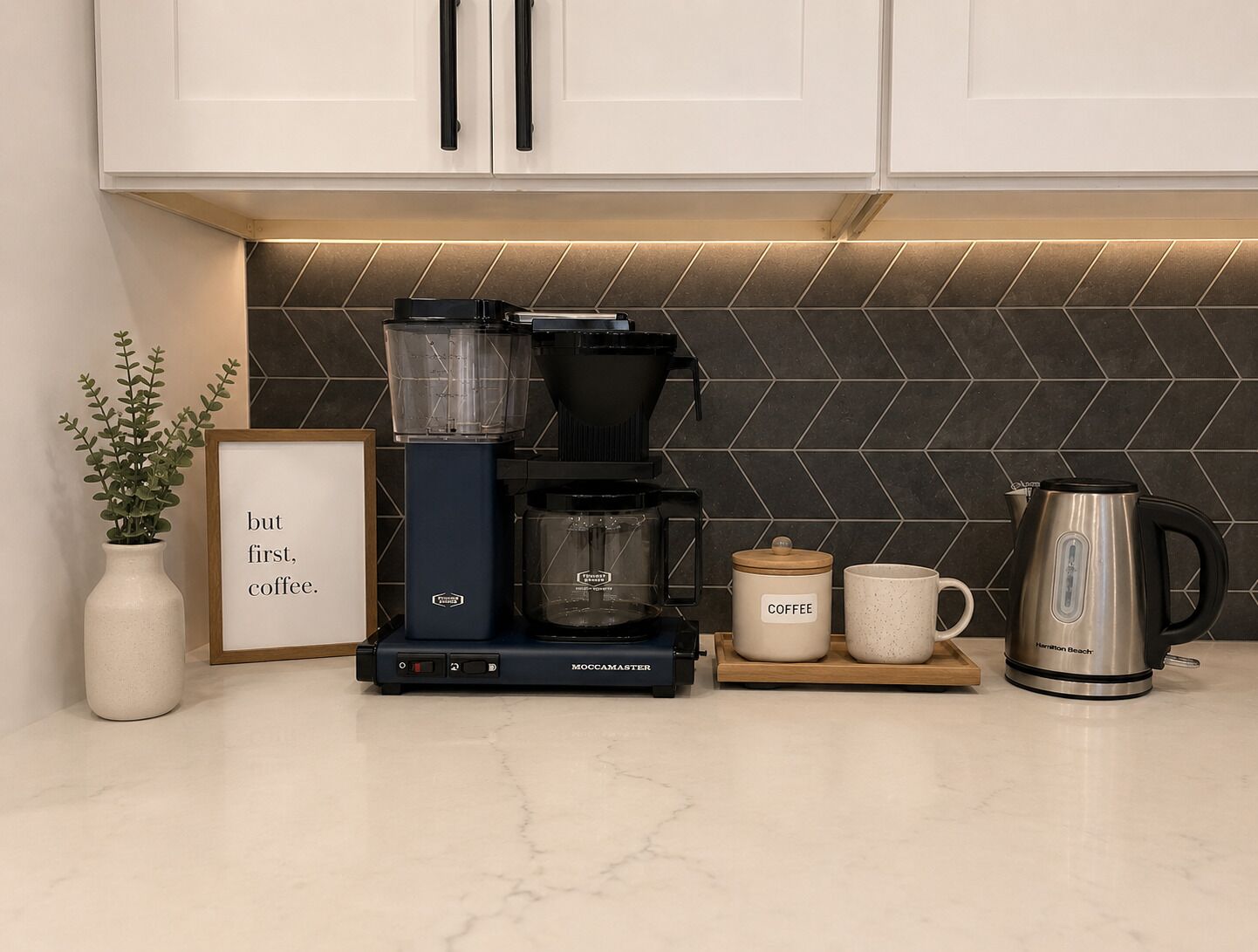A kitchen with a coffee maker, two coffee cups, a kettle, and a framed sign.