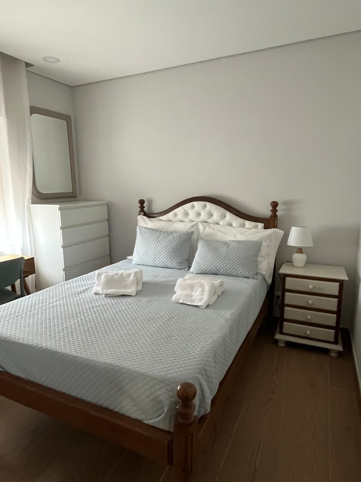 A neatly made bed with a wooden headboard, a bedside table with a lamp, and a chair in a room with a mirror on the wall.