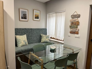 A living room with a glass dining table, green chairs, a grey sofa with patterned cushions, a window with white curtains, and two framed pictures on the wall. - Coral House (Viana do Castelo)