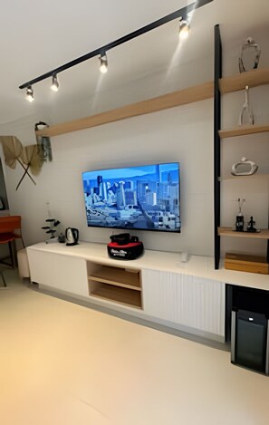 A modern living room with a flat-screen TV mounted on a white entertainment unit, wooden shelves above, and a small black appliance below. - Casa Dez Guarajuba  (Camaçari)