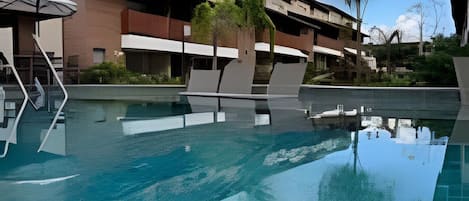 A swimming pool with lounge chairs, a covered seating area, and a building with multiple balconies in the background.