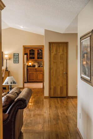A hallway with a wooden floor, a brown leather sofa, a wooden door, and a kitchen in the background. - Lakepoint Overlook | Dock + Lake Access (Bella Vista)