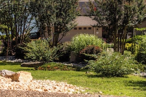 A garden with various plants and trees, a building with a beige facade, and a clear sky. - Lakepoint Overlook | Dock + Lake Access (Bella Vista)