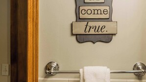A bathroom with a towel rack, a wall-mounted sign with the phrase 'Dreams do come true.', and a light switch. - Lakepoint Overlook | Dock + Lake Access (Bella Vista)