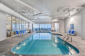 An indoor swimming pool with clear blue water, a view of the sea through large windows, and blue chairs along the poolside. - Admirals Quarters 1201 (Orange Beach)