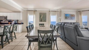 A spacious living area with a dining table, chairs, a gray sofa, a kitchen with white cabinets, and a view of the ocean. - Admirals Quarters 1201 (Orange Beach)