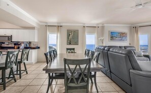 A spacious living area with a dining table, chairs, a gray sofa, a kitchen with white cabinets, and a view of the ocean. - Admirals Quarters 1201 (Orange Beach)