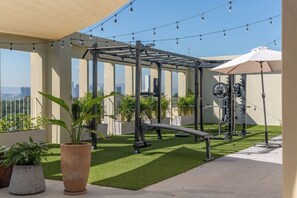 Zona de gimnasio al aire libre con equipo de ejercicio, plantas en macetas y vistas a la ciudad.