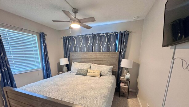 A bedroom with a wooden bed, a ceiling fan, a TV mounted on the wall, and a window with blinds. - Welcome to Your Beachside Escape Tucked Away on the Serene Fort Morgan Peninsula (Gulf Shores)