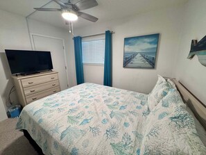 Une chambre à coucher avec un lit, un ventilateur de plafond, un téléviseur posé sur une commode, une fenêtre avec des rideaux et un tableau accroché au mur.