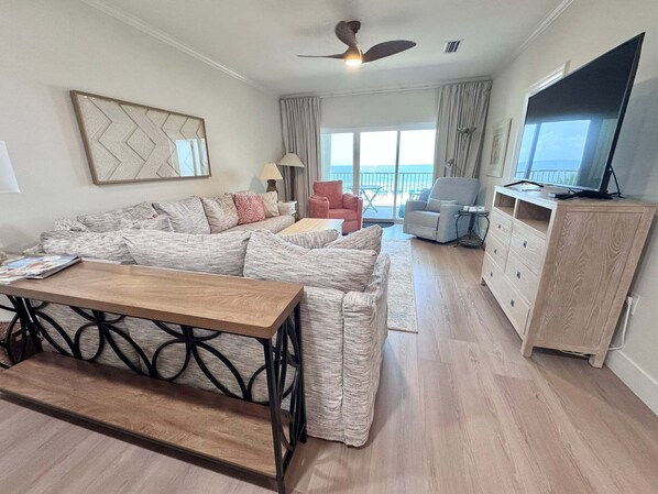 Uma sala de estar com sofá, mesa de centro, televisão e varanda com cortinas.