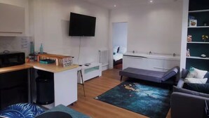 Een ruimtelijk ingerichte leefruimte met kitchenette, een wandtelevisie, een bank en een kleine eettafel.