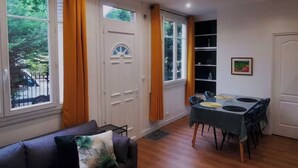 Een kamer met een eettafel, stoelen, een bank en grote ramen voorzien van gordijnen.