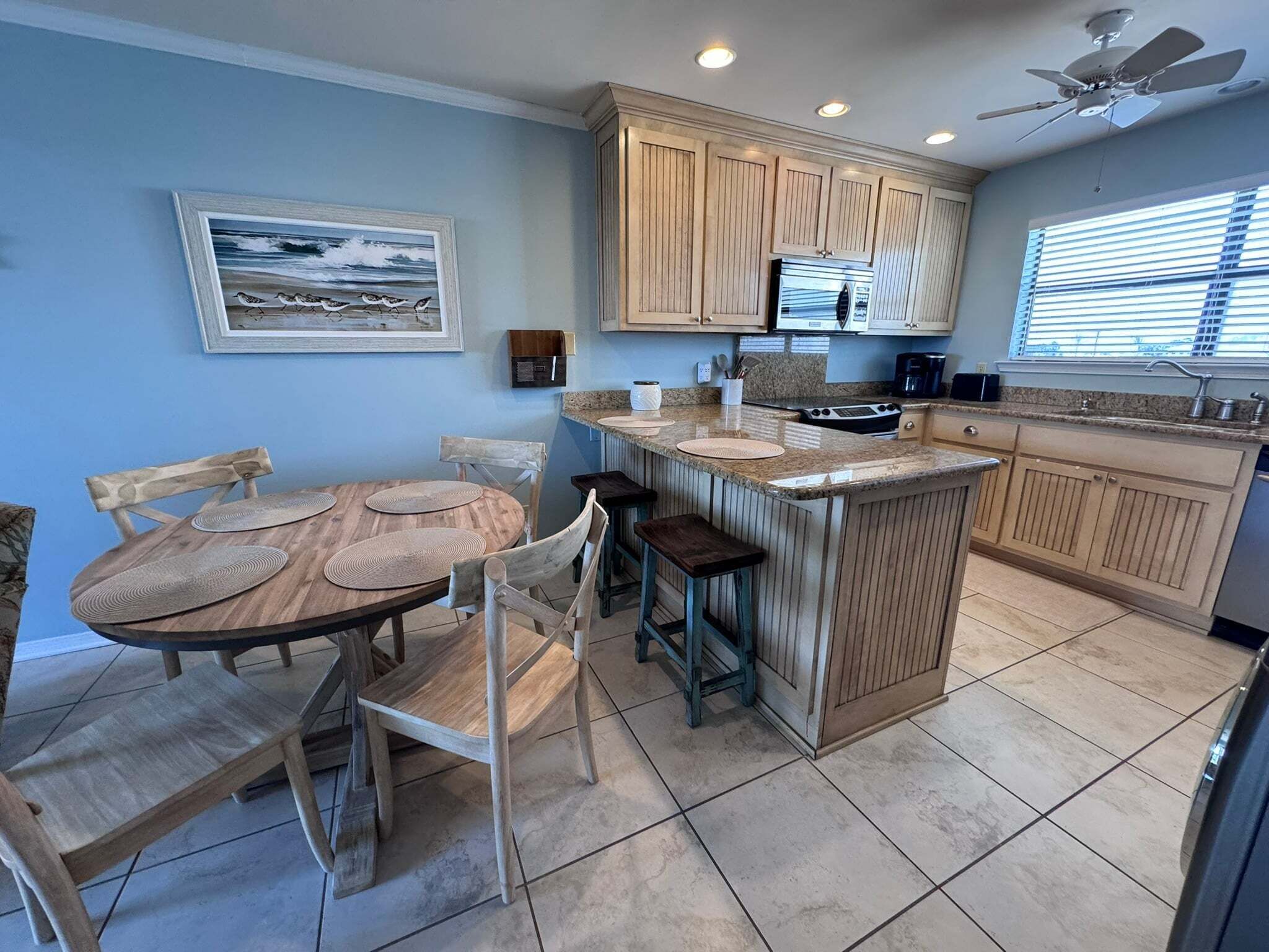 A kitchen with wooden cabinets, a granite countertop, a dining area with a wooden table and chairs, and a ceiling fan.