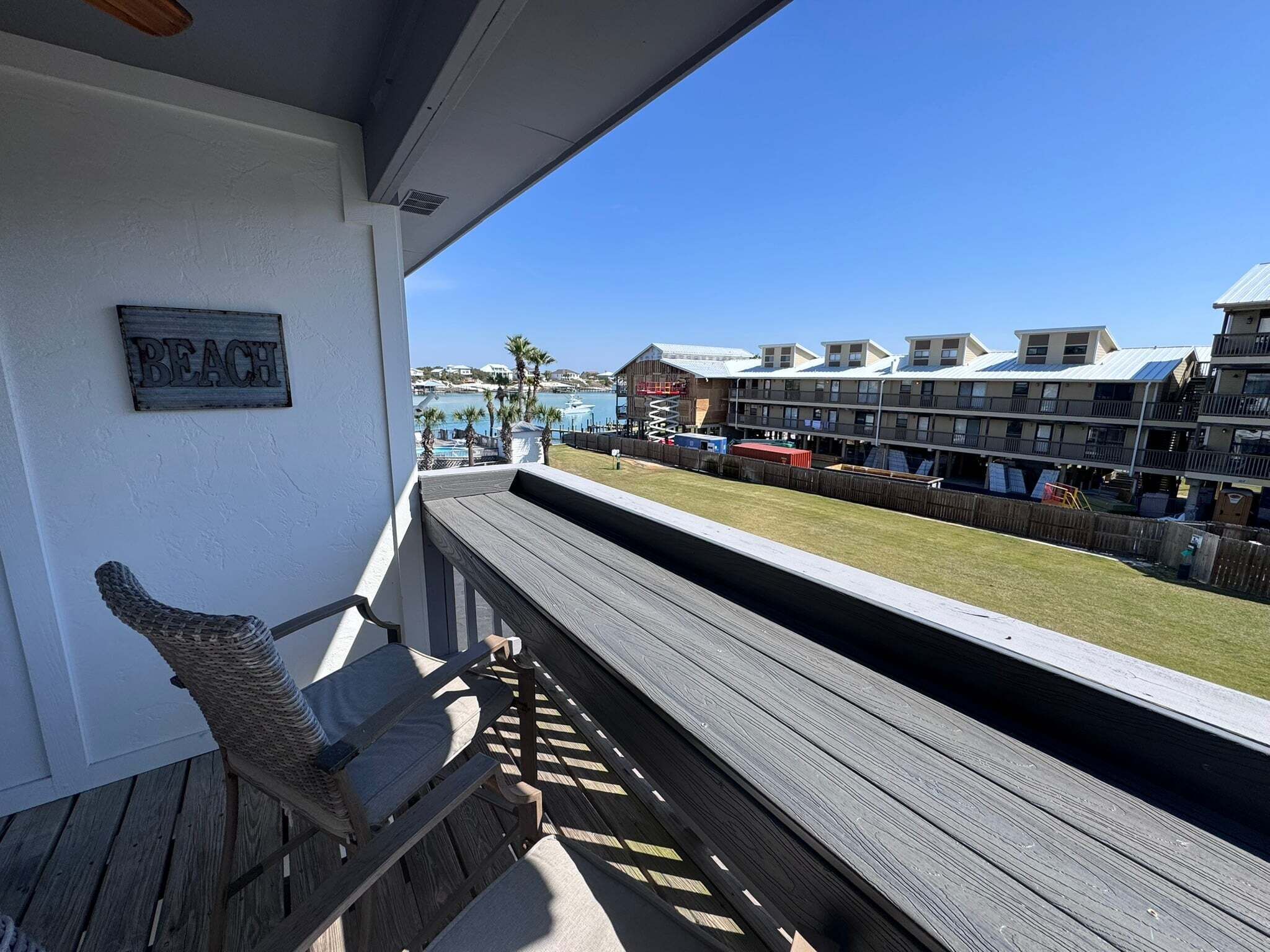 A balcony with a wicker chair, a view of a beach, and a row of buildings in the background.