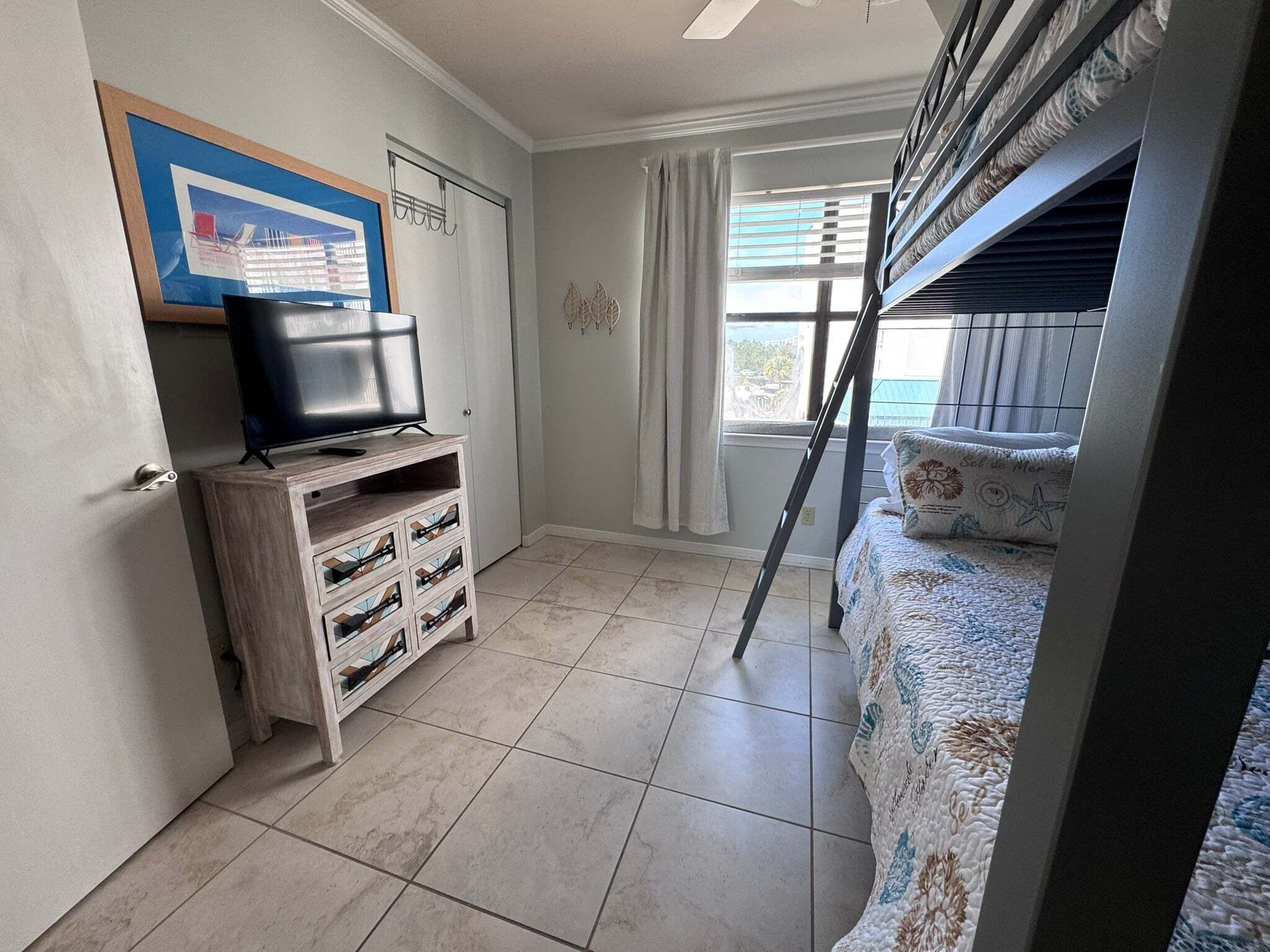 Une chambre avec un lit superposé, un téléviseur posé sur un meuble en bois, une fenêtre avec des rideaux et un tableau encadré accroché au mur.
