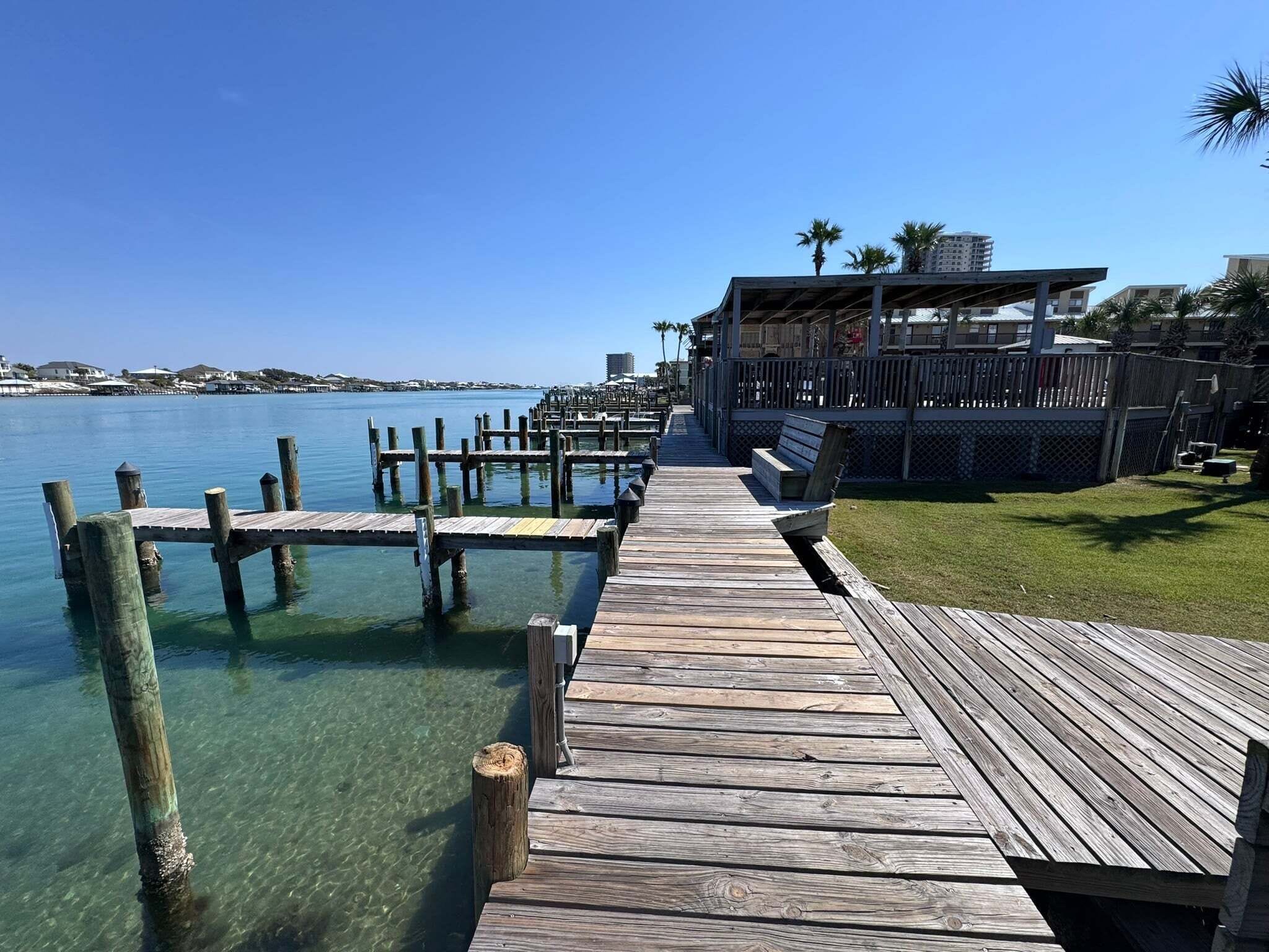 A wooden pier leading to a dock with clear water, a building with a covered area, and palm trees in the background.