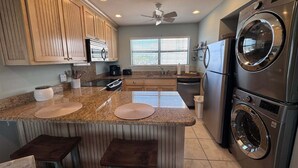A kitchen with granite countertops, wooden cabinets, a built-in oven, a refrigerator, and a wall-mounted washer and dryer. - Coastal Comfort w/ Pool + Easy Beach Access 2br/2ba (Orange Beach)