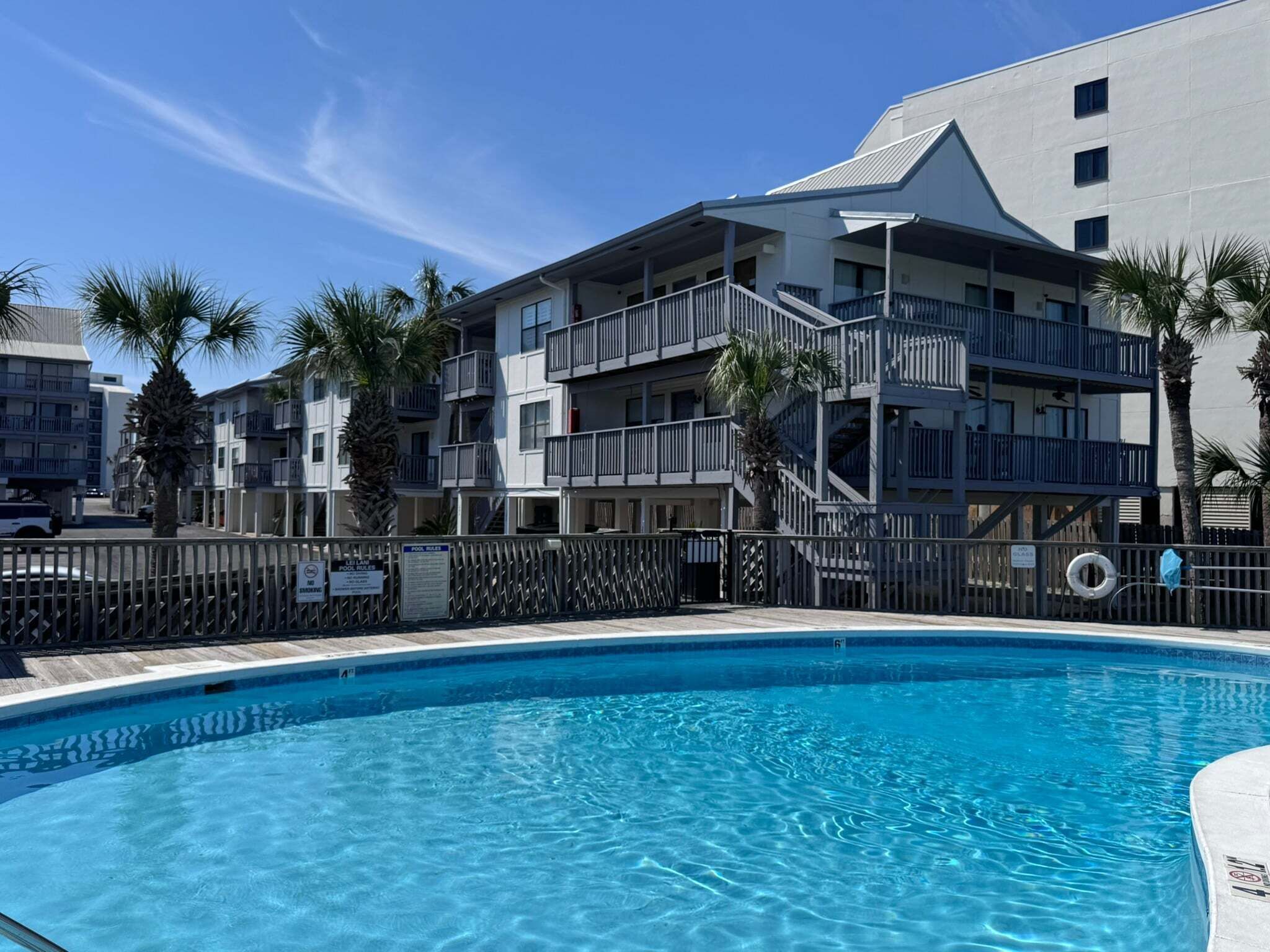 A swimming pool with a clear blue water, surrounded by a fence. In the background, there are multi-story buildings with balconies and palm trees.