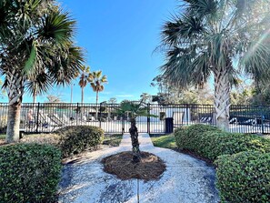 A pathway leading to a pool area, surrounded by palm trees and a fence. - Welcome to This 2br/2ba Relaxing Coastal Escape (Orange Beach)