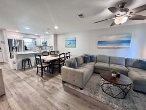 A modern living room with a sectional sofa, a dining area with a table and chairs, and a kitchen with white cabinets and appliances. - Welcome to This 2br/2ba Relaxing Coastal Escape (Orange Beach)