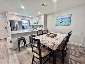 A dining area with a rustic table, wooden chairs, and a kitchen in the background. - Welcome to This 2br/2ba Relaxing Coastal Escape (Orange Beach)