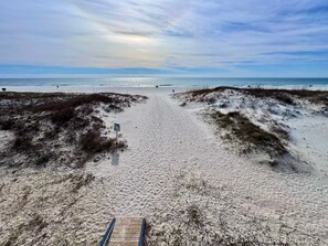Ein Sandstrand mit einer Holzpromenade, die zum Wasser führt, einem Wegweiser und Dünen.