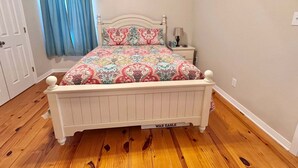 A bedroom with a white bed, patterned bedding, a nightstand with a lamp, and a window with blue curtains. - Welcome to This 3br/2ba Charming Coastal Cottage (Gulf Shores)