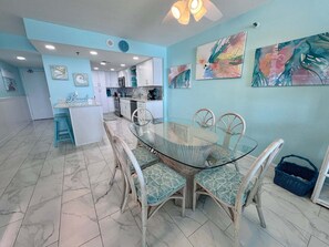 A dining area with a glass table and wicker chairs, a kitchen in the background, and abstract art on the walls. - Welcome to a Beautifully Updated 2br/2ba Gulf-front Escape (Orange Beach)