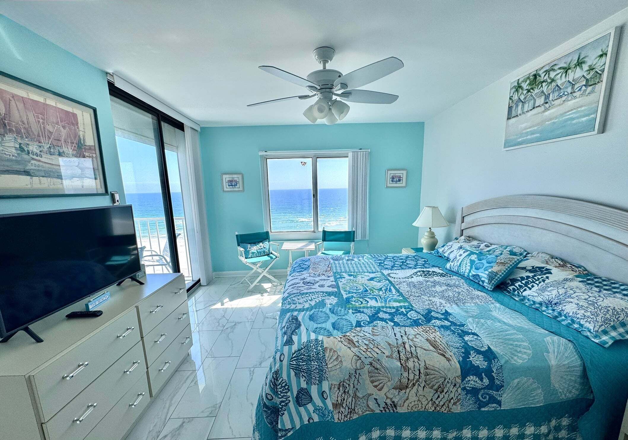 A bedroom with a bed, a TV, a ceiling fan, a window with a view of the ocean, and a chair.