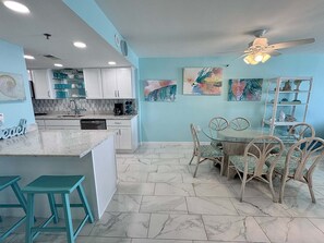 A modern kitchen with white cabinets, a central island, and a dining area with a glass table and wicker chairs. - Welcome to a Beautifully Updated 2br/2ba Gulf-front Escape (Orange Beach)