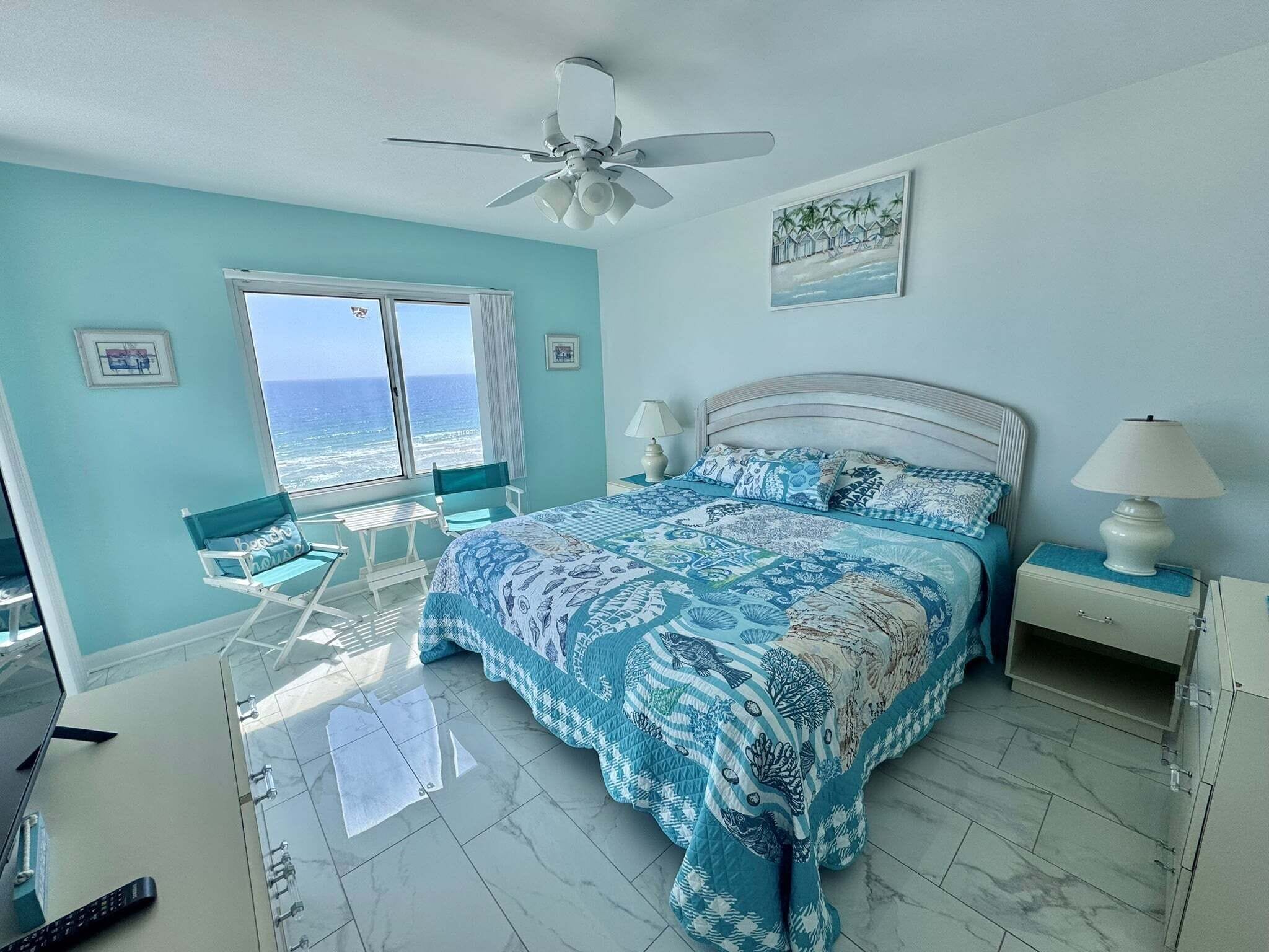 A bedroom with a large bed, a ceiling fan, a view of the ocean, and a beach scene painting.