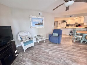A living room with a television, wicker chairs, a dining table, and a kitchen in the background. - Gulf Shores Getaway! 2BR Pool Walk to Beach Pet Friendly (Gulf Shores)