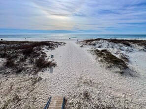 Ein Sandstrand mit einer Holzpromenade, die zum Wasser führt, einem Wegweiser und Dünen.