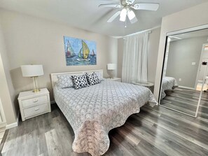 A bedroom with a bed, nightstands, a ceiling fan, a painting, and a mirror. - Gulf Shores Getaway! 2BR Pool Walk to Beach Pet Friendly (Gulf Shores)