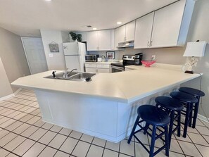 A modern kitchen with a central island, bar stools, and a microwave. - Gulf Shores Getaway! 2BR Pool Walk to Beach Pet Friendly (Gulf Shores)